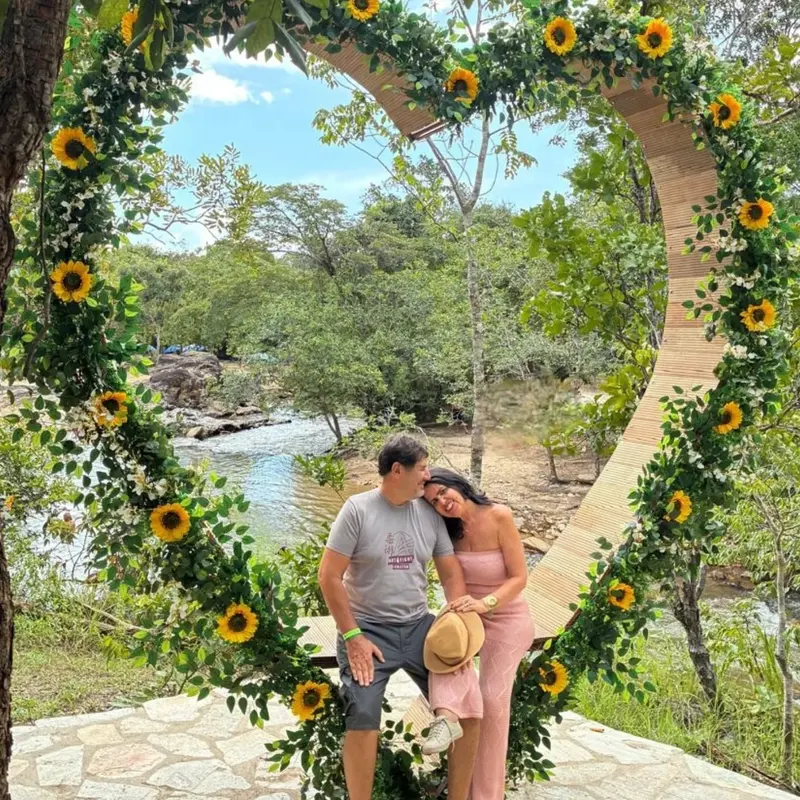 Casal dentro de um arco em forma de coração coberto de girassóis, com o rio ao fundo.