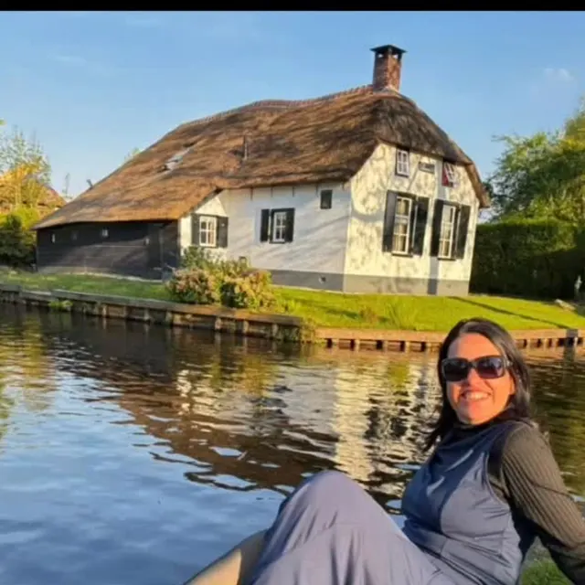 Viajante sentada à beira de um canal em Giethoorn, com uma casa de telhado de palha ao fundo.