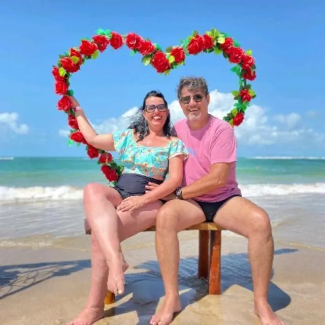 O casal sentado num banco na areia, dentro de uma moldura de coração com flores.