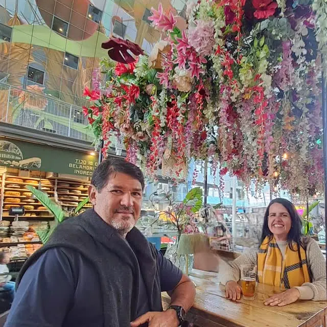 O casal numa mesa do Markthal, sob flores e frutas penduradas.